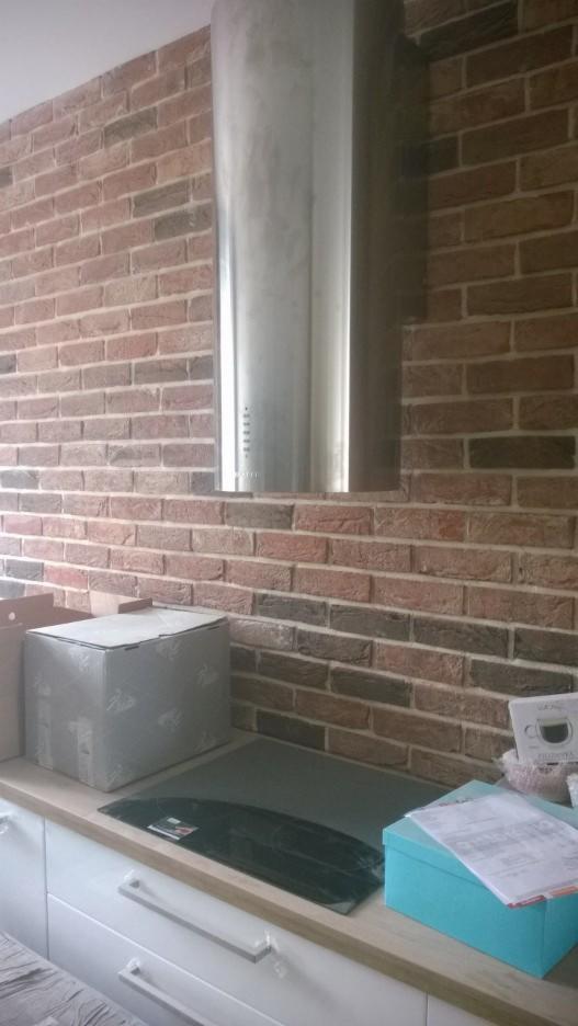 Kuchnia po remoncie: ceglana ściana, metalowy okap i białe szafki. Widoczne kartony i akcesoria kuchenne na blacie. Styl rustykalny z nowoczesnymi elementami.