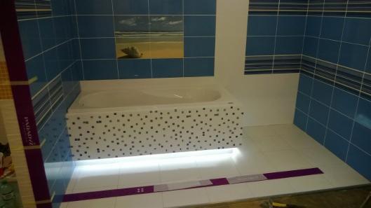 Wykończona łazienka z niebieskimi płytkami, mozaikową obudową wanny i podświetleniem LED. Na ścianie dekoracja z motywem morskim. Widoczne materiały wykończeniowe.