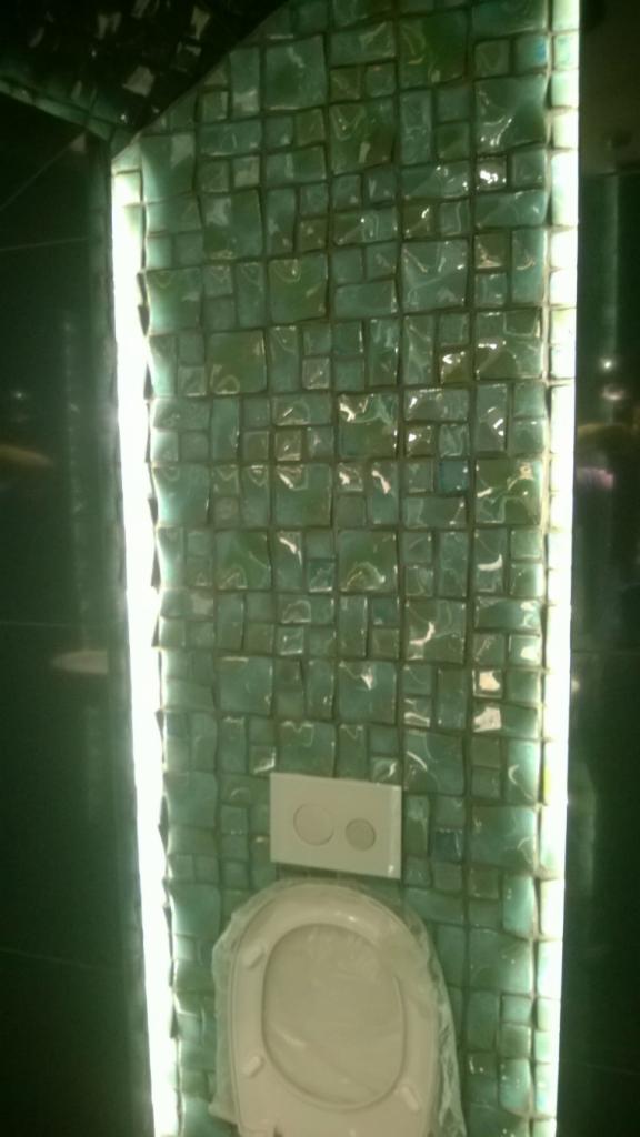 Nietypowa ściana w łazience z zielonej mozaiki, podświetlona po bokach. Widoczny nowy sedes w folii ochronnej i przycisk spłuczki. Całość tworzy nowoczesny wystrój.