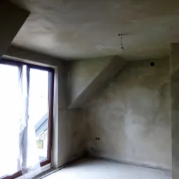 Pokój na poddaszu po nałożeniu gładzi, widoczne surowe wykończenie, okno z zasłoniętą szybą, wystające kable oświetleniowe i gniazdka elektryczne.