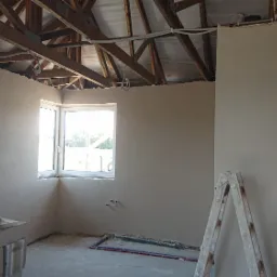 Wnętrze w trakcie prac wykończeniowych: świeżo otynkowane ściany w szarym odcieniu, drewniana więźba dachowa, okno narożne z widokiem na zieleń, para butów sportowych i robocza bluza leżące...