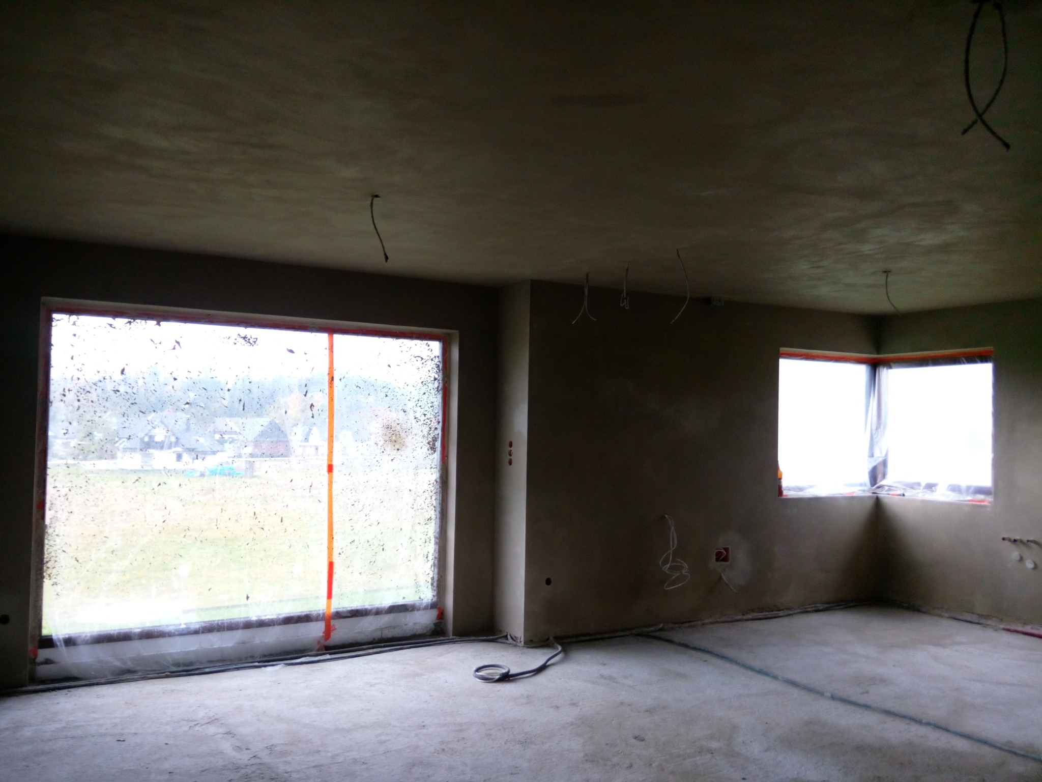 Surowe wnętrze w trakcie prac wykończeniowych, ściany pokryte świeżym tynkiem, okna zabezpieczone folią, widoczne przewody elektryczne.