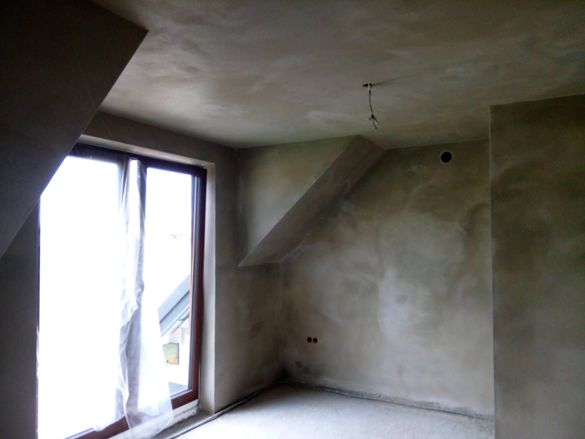 Pokój na poddaszu po nałożeniu gładzi, widoczne surowe wykończenie, okno z zasłoniętą szybą, wystające kable oświetleniowe i gniazdka elektryczne.