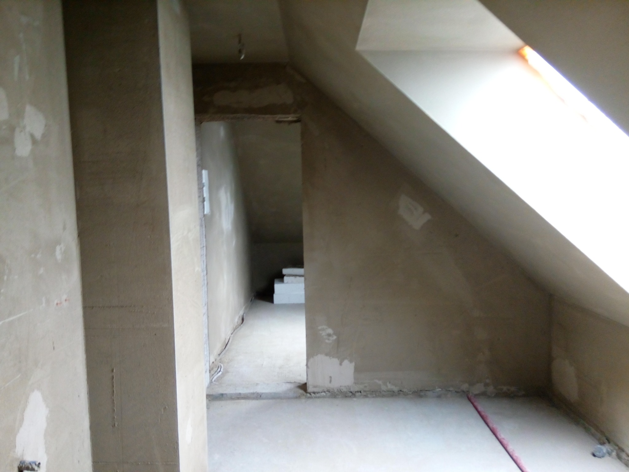 Surowe wnętrze poddasza w trakcie prac wykończeniowych, widoczne ściany pokryte świeżym tynkiem, przygotowane do dalszych etapów prac, z oknem dachowym wpuszczającym jasne światło.