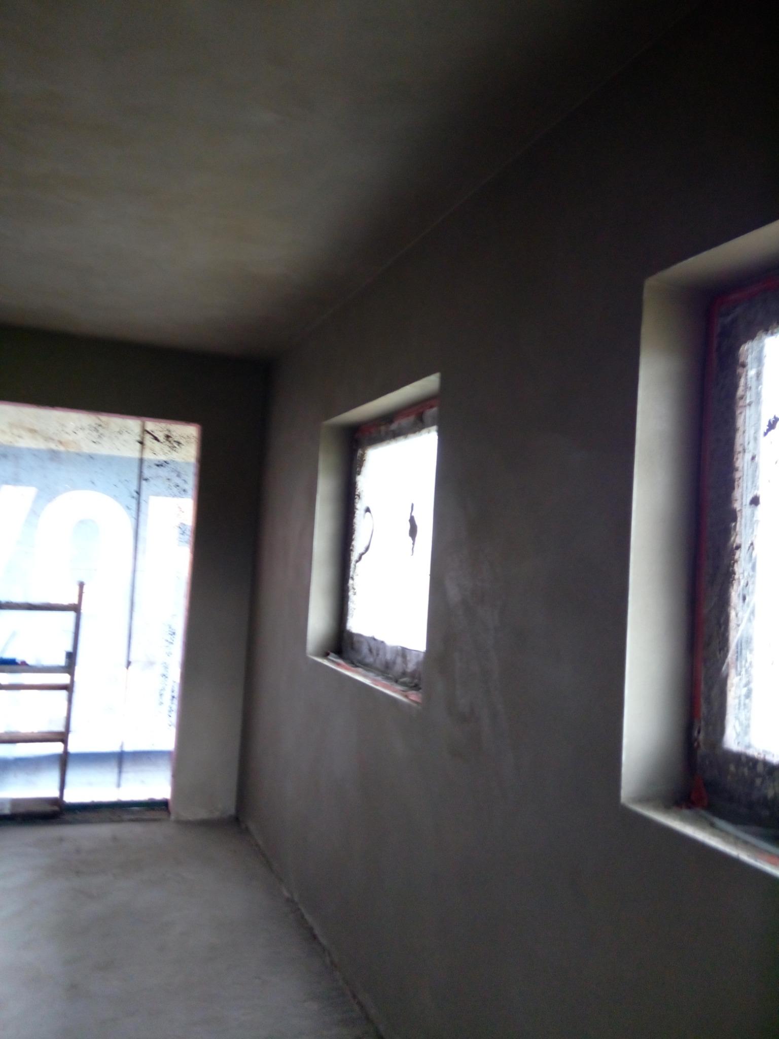 Świeżo otynkowane ściany wewnętrzne w pomieszczeniu, widoczne dwa okna z zabezpieczeniami i drabina oparta o ścianę przy wejściu, surowy stan wykończenia.