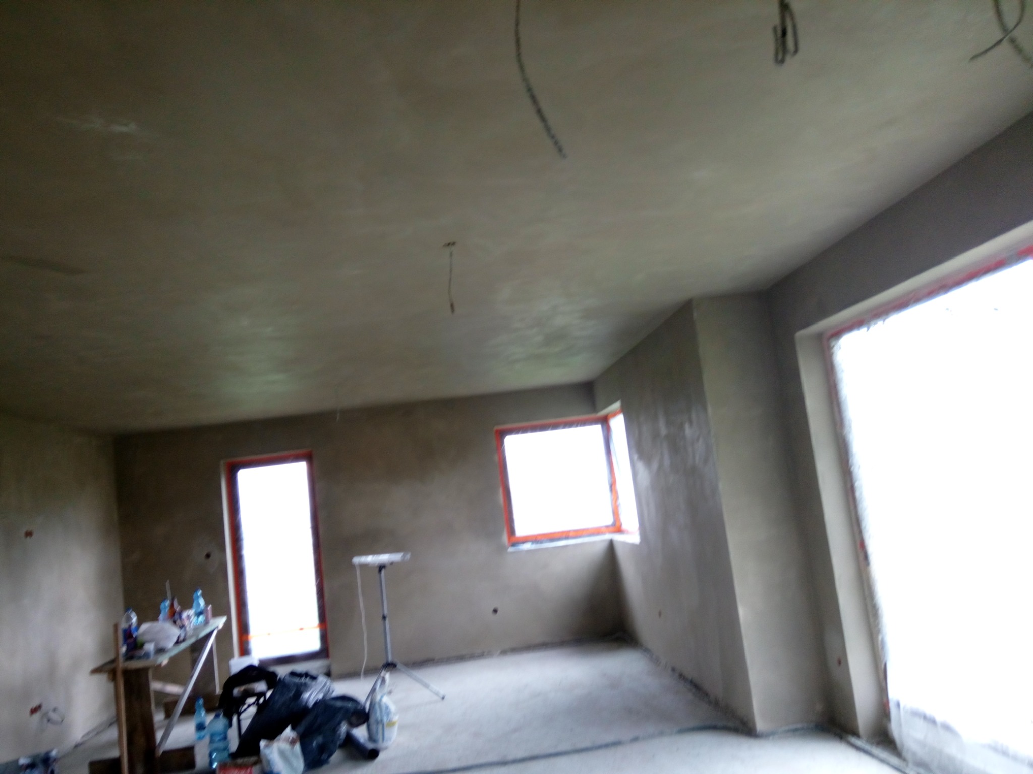 Pokój w trakcie prac wykończeniowych, ściany i sufit pokryte świeżym tynkiem, widoczne okna z pomarańczowymi ramami i prowizoryczny stół z butelkami wody.