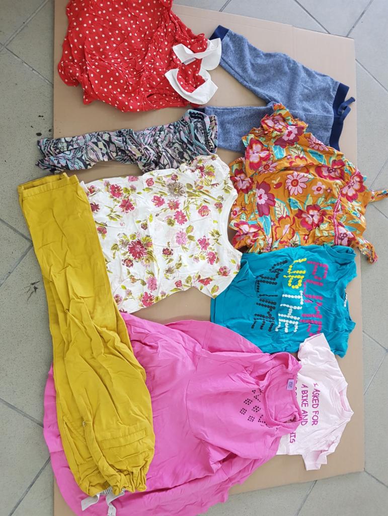 Stos ubrań używanych na kartonie: czerwona sukienka w białe kropki, żółte spodnie, koszulki z nadrukami i bluzki w kwiatowe wzory.