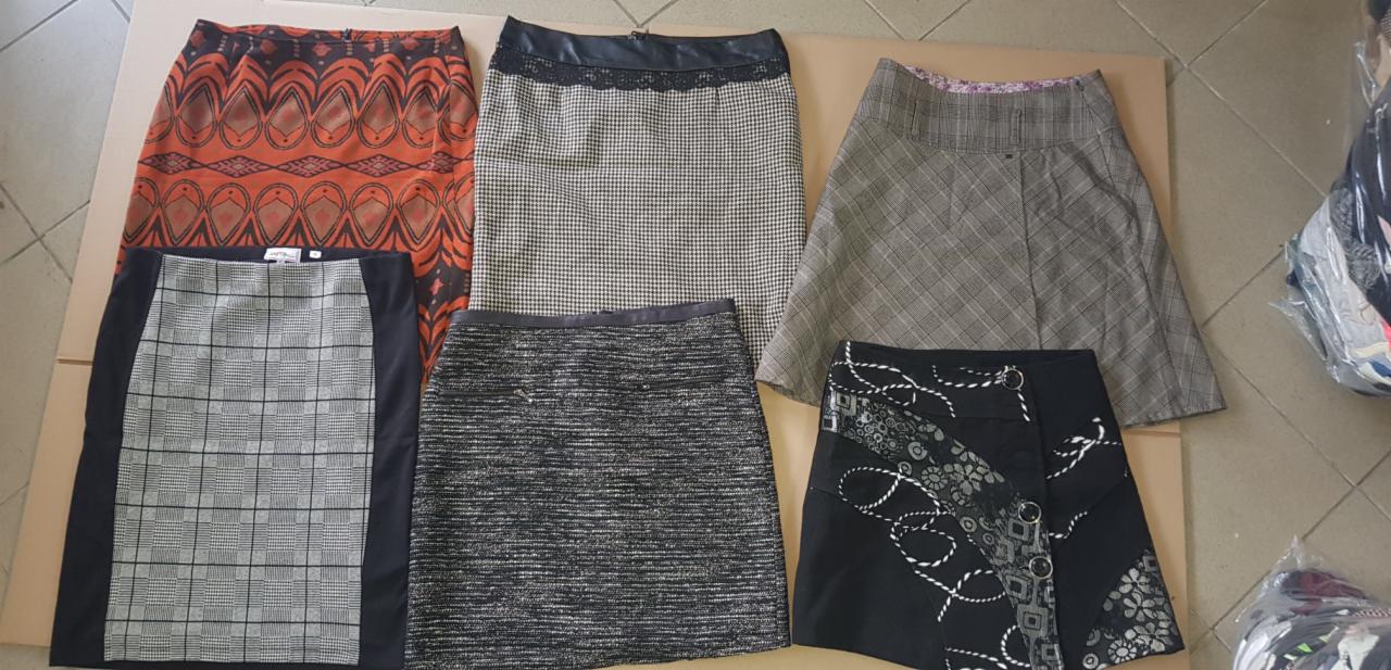 Zestaw używanych spódnic o różnych wzorach i długościach, oferowanych w Warszawie, ułożonych na kartonowym podłożu. Widoczna spódnica w pomarańczowe wzory, krótka spódnica w kratę, spódnica...