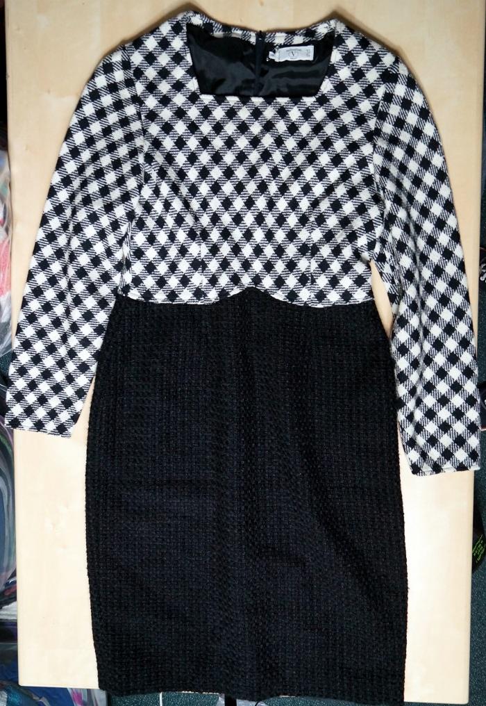 Czarno-biała sukienka w kratę z długim rękawem i czarnym dołem, leżąca na drewnianej powierzchni. Widoczny zamek błyskawiczny i metka.