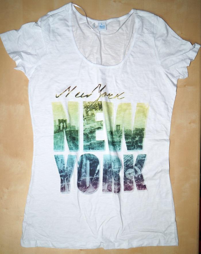 Biała damska koszulka z krótkim rękawem, z nadrukiem 'New York' w kolorach gradientu i motywem miasta wewnątrz liter, leżąca na drewnianym podłożu.