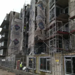 Wielopiętrowy budynek w trakcie prac elewacyjnych, z rusztowaniami i osłonami z logo Atlas. Widoczni pracownicy w odblaskowych kamizelkach. Częściowo ogrodzony teren budowy.