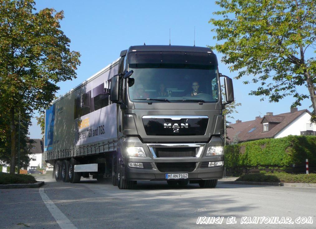 Szary ciągnik MAN TGX z naczepą na drodze, z widocznymi osobami w kabinie, drzewami i budynkiem w tle. Tablica rejestracyjna MAN 7102.
