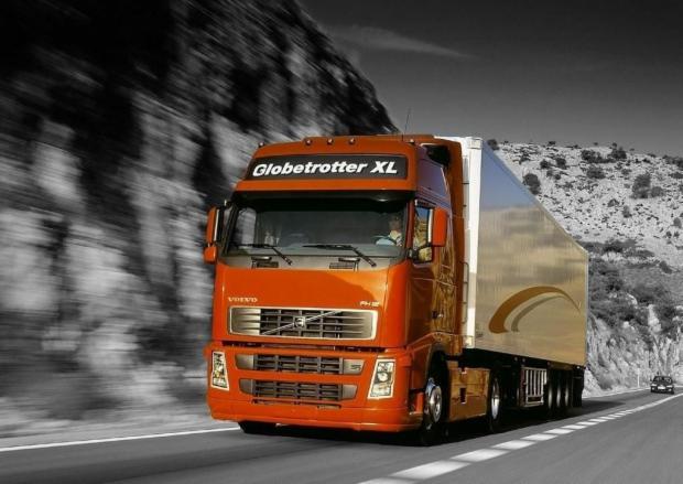 Pomarańczowa ciężarówka Volvo Globetrotter XL w ruchu na drodze, z rozmytym tłem górskim. Widoczny kierowca i fragment naczepy. Dynamiczne ujęcie.