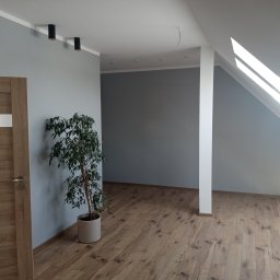 BUDOPLEX-ADAM BUDNIK - Pomalowane na szaro wnętrze poddasza z drewnianą podłogą, oświetleniem punktowym i rośliną w doniczce, widok na sąsiednie pomieszczenie przez otwartą przestrzeń.