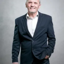 Portret uśmiechniętego, starszego mężczyzny w granatowej marynarce i dżinsach, stojącego na tle szarego tła. Lewa ręka w kieszeni, widoczny zegarek.