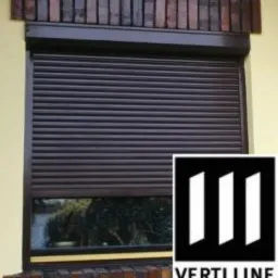 Ciemnobrązowa, zwinięta osłona okienna z logo Verti Line w prawym dolnym rogu, zamontowana na elewacji budynku z ceglaną dekoracją nad oknem.