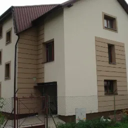 Dwukondygnacyjny dom z beżową elewacją i brązowymi akcentami wokół okien oraz poziomymi pasami. Widoczne ogrodzenie z siatki i roślinność.