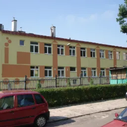 Dwukondygnacyjny budynek z żółtą elewacją i pomarańczowymi akcentami, widoczny zza ogrodzenia. W tle drzewo, a na pierwszym planie zaparkowane samochody.