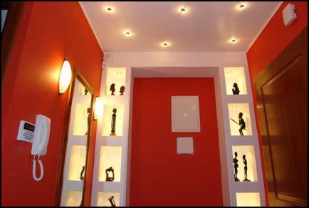 Czerwone ściany holu z wnękami oświetlonymi punktowo, w których umieszczono dekoracyjne figurki. Na ścianie wisi lustro i telefon.