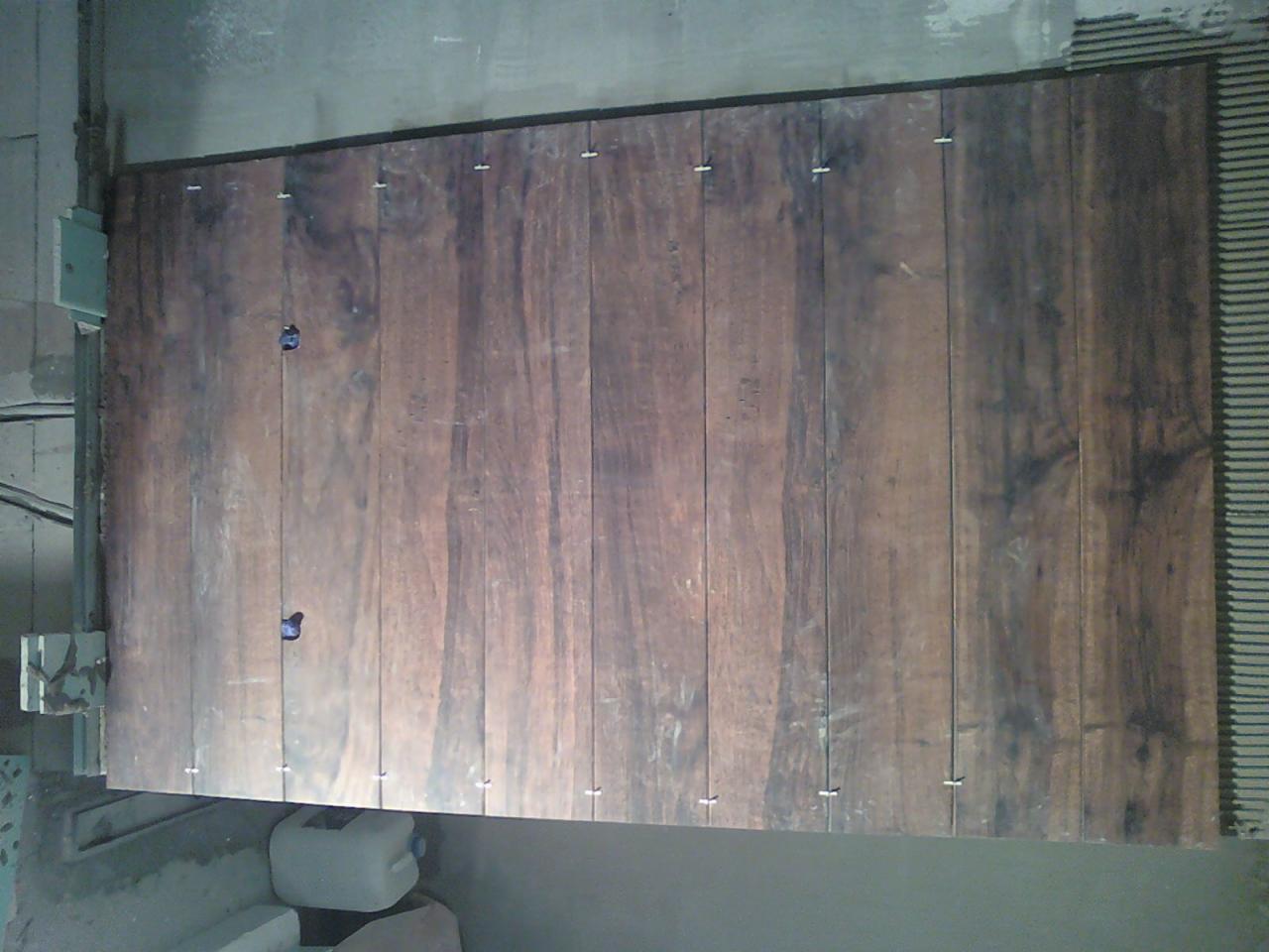 Ułożone płytki imitujące drewno, z krzyżykami dystansowymi, na ścianie pokrytej klejem. Widoczny fragment narzędzia do cięcia płytek.