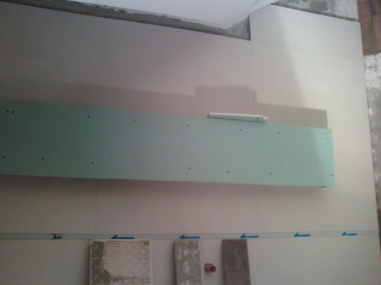 Montaż płyty gipsowo-kartonowej na ścianie z położoną częściowo glazurą. Widoczne ślady kleju i poziomica. Prace wykończeniowe w łazience.