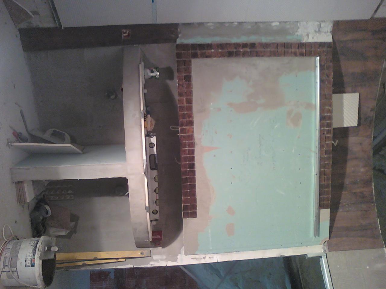 Wnętrze w trakcie remontu: częściowo wykończona ściana z brązową mozaiką, surowe półki, wiadro z zaprawą. Widoczny poziomica i inne narzędzia. Prace wykończeniowe.