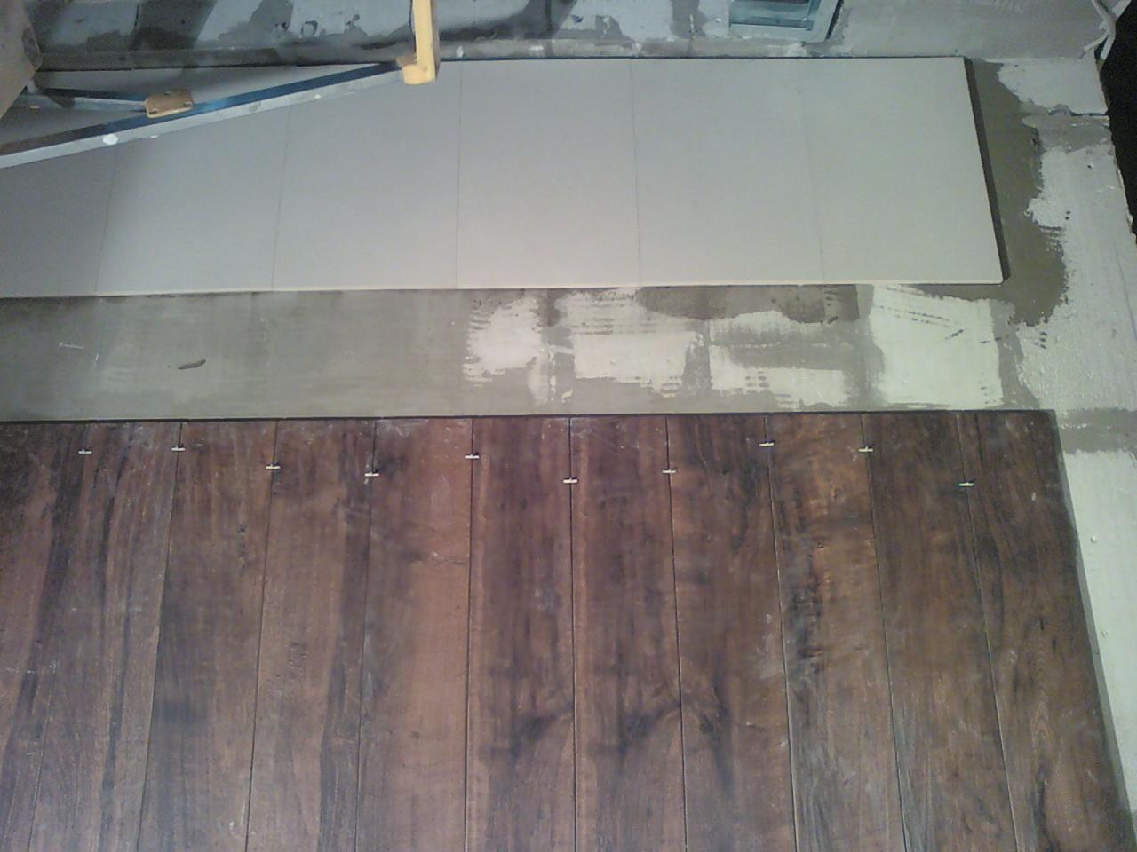 Układanie paneli podłogowych w odcieniach brązu, widoczne kliny dystansowe. Częściowo ułożone, widoczny fragment podkładu i surowy beton. Ujęcie z góry.