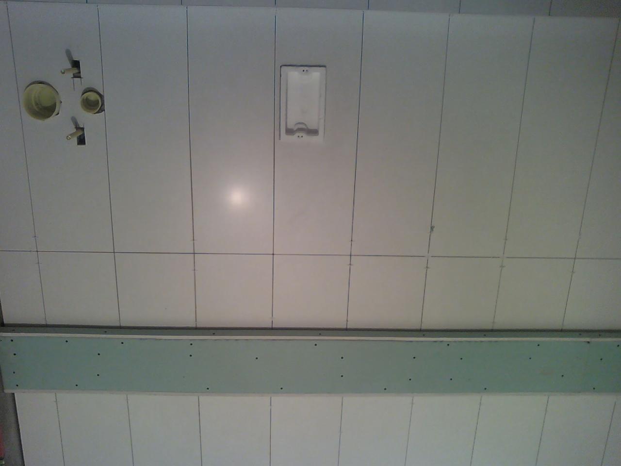 Białe płytki ścienne z widocznymi otworami na instalacje i zielonym profilem montażowym. Ujęcie z dołu, podkreślające precyzję układania płytek.