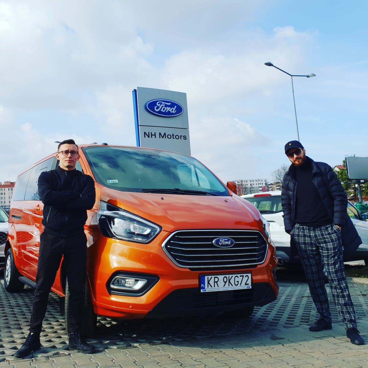 Pomarańczowy Ford Transit z dwoma mężczyznami na tle salonu samochodowego NH Motors; tablica rejestracyjna KR 9KG72.