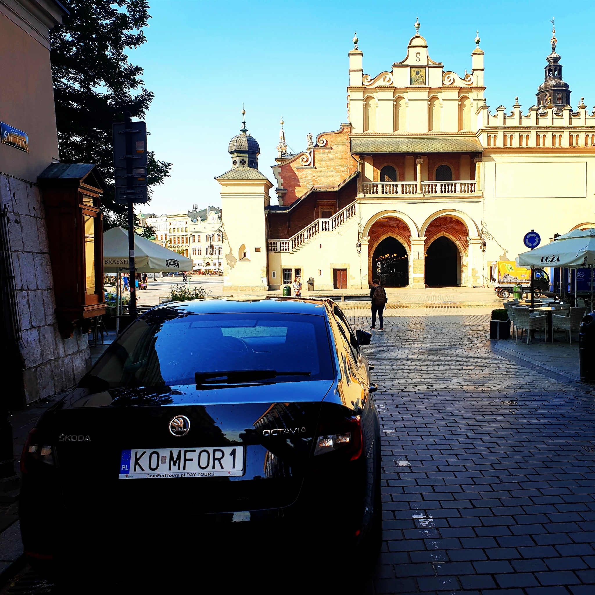 Czarny samochód Skoda Octavia z tablicą rejestracyjną 'KO MFOR1' zaparkowany na brukowanej ulicy w Krakowie, na tle Sukiennic i placu z parasolami kawiarnianymi.