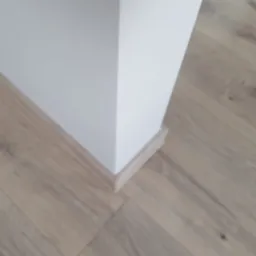 Fragment rogu pomieszczenia z ułożonymi panelami podłogowymi w odcieniach beżu i dopasowaną listwą przypodłogową, ściana pomalowana na biało.