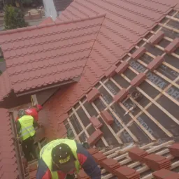 Montaż czerwonej dachówki ceramicznej na drewnianej konstrukcji dachu, widoczni dekarze w odblaskowych kamizelkach, częściowo ukończony fragment pokrycia dachu z widocznym deskowaniem i membraną...