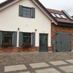 Nowoczesny dom z białymi ścianami, ceglaną elewacją do połowy wysokości, szarymi okiennicami i garażem, z kwiatami w donicach pod oknami i brukowanym podjazdem.