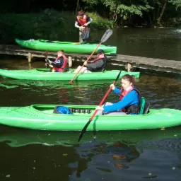 Grupa osób płynących na zielonych kajakach po spokojnej rzece w słoneczny dzień. Drewniany pomost i zielona roślinność w tle.