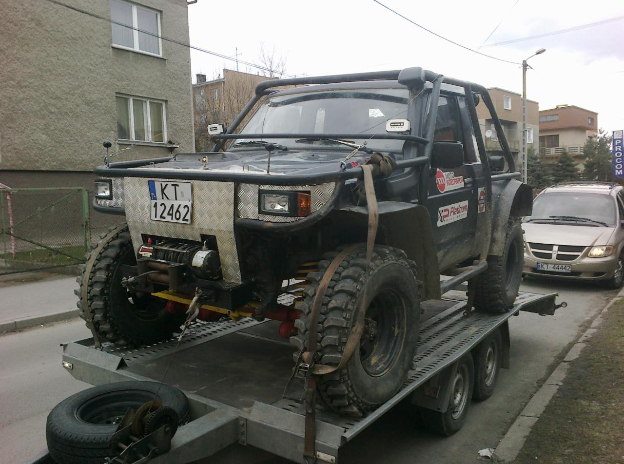 Czarny, zmodyfikowany samochód terenowy z wyciągarką, przewożony na niskopodwoziowej lawecie. Widoczny fragment ulicy i budynki w tle. Tablica rejestracyjna KT 12462.