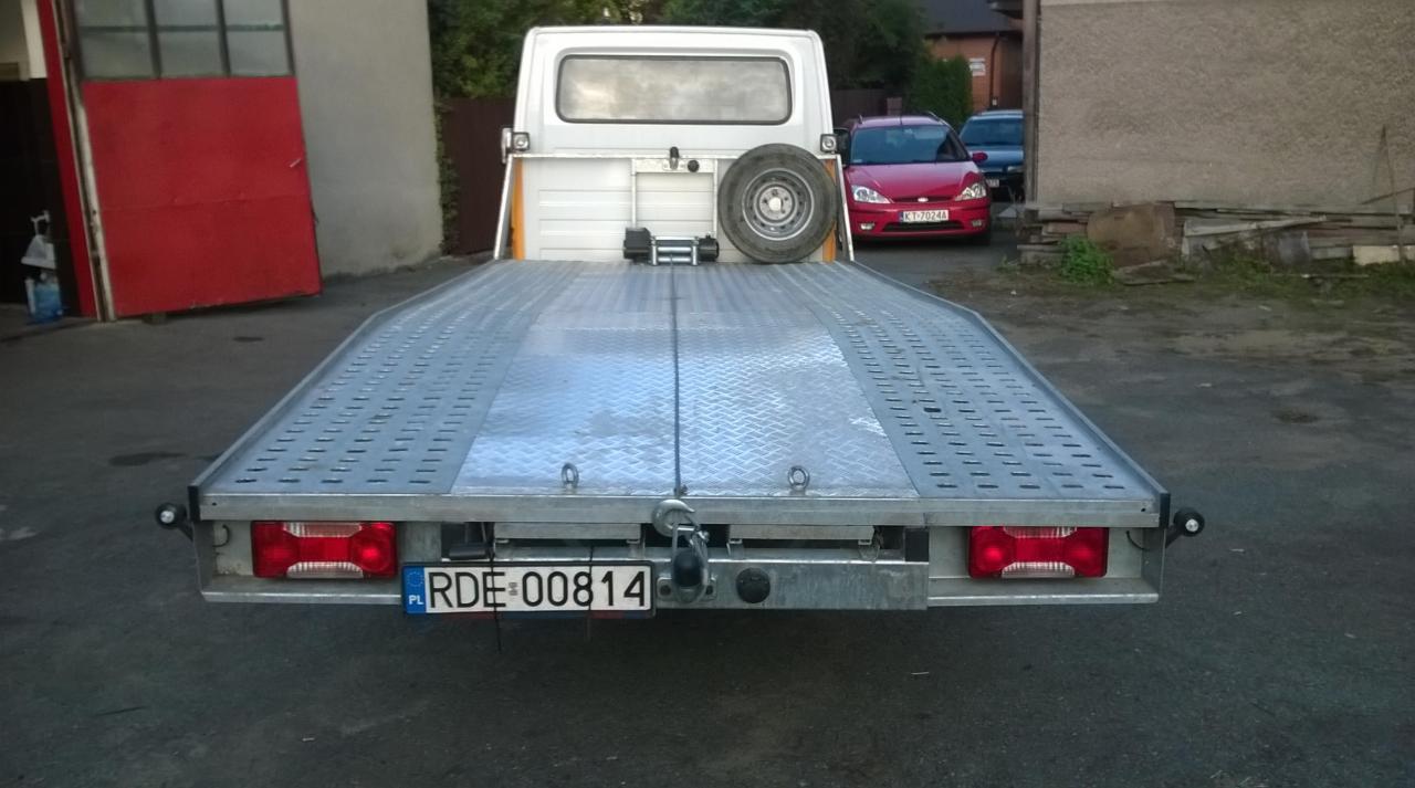 Widok z tyłu lawety z podnośnikiem, na której znajduje się koło zapasowe i wciągarka. W tle zaparkowane samochody i budynek.