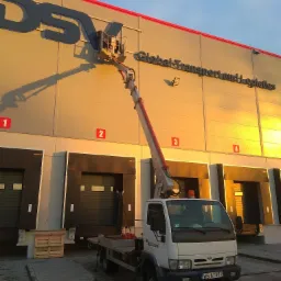Podnośnik koszowy z pracownikiem na wysokości podczas montażu lub czyszczenia dużego logo firmy 'DSV Global Transport and Logistics' na elewacji magazynu z rampami załadunkowymi w słoneczny dzień.