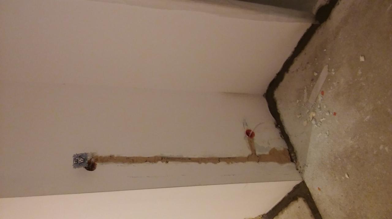 Ściana z wyżłobionymi bruzdami pod instalację elektryczną, widoczne puszki instalacyjne i przewody w czerwonej osłonie, surowy beton na podłodze.