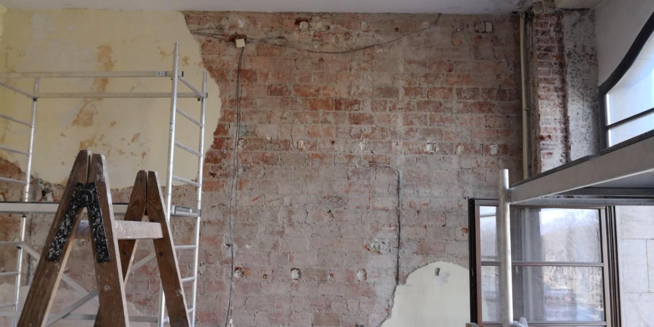 Odsłonięta ceglana ściana z fragmentami starego tynku, widoczne przewody elektryczne, drewniana drabina i aluminiowe rusztowanie.