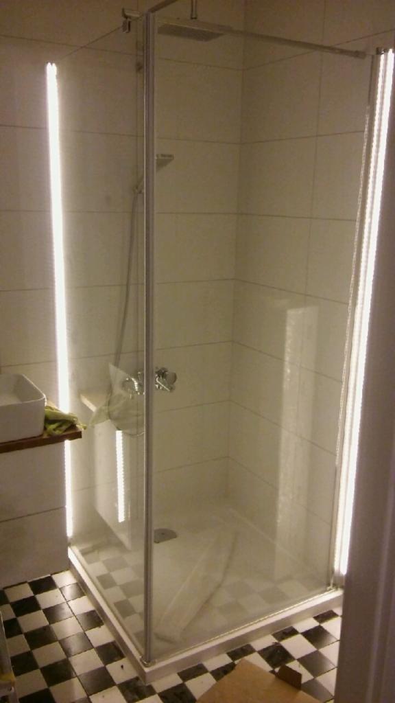 Nowoczesna kabina prysznicowa z deszczownicą, szklanymi ścianami i czarno-białą podłogą w szachownicę, oświetlona pionowymi lampami LED.