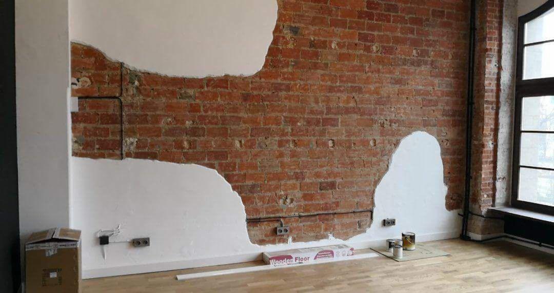 Częściowo otynkowana ceglana ściana z widocznymi przewodami elektrycznymi, drewniana podłoga, puszki z farbą i karton z napisem 'Wooden Floor'.