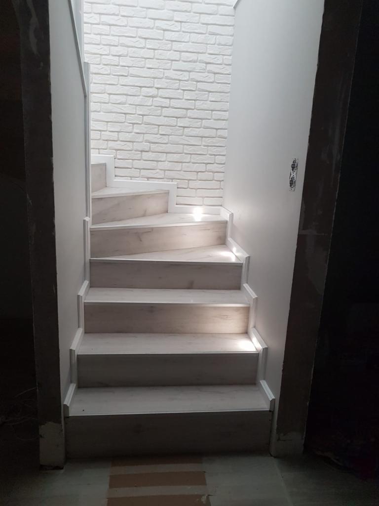 Schody wewnętrzne z jasnego drewna, prowadzące do góry, ściana wyłożona białą cegłą dekoracyjną, widoczne niedokończone wykończenia.