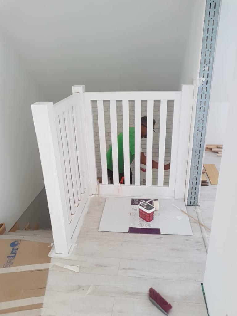 Biała, drewniana barierka schodowa z pionowymi szczeblami w trakcie montażu, zabezpieczająca dostęp do schodów. W tle widoczna osoba w zielonej koszulce. Na podłodze kartony, farba i pędzel