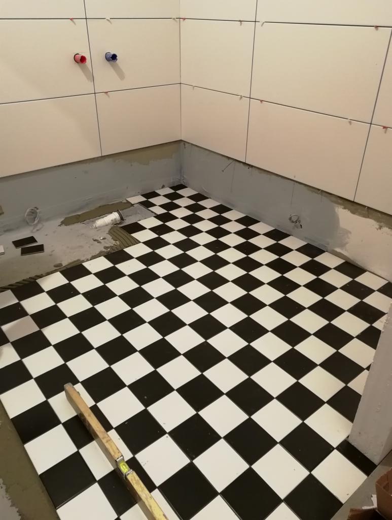Układanie czarno-białej szachownicy z płytek na podłodze w łazience, poziomnica leży na ułożonej powierzchni, widoczne przygotowane ściany i wystające rury.