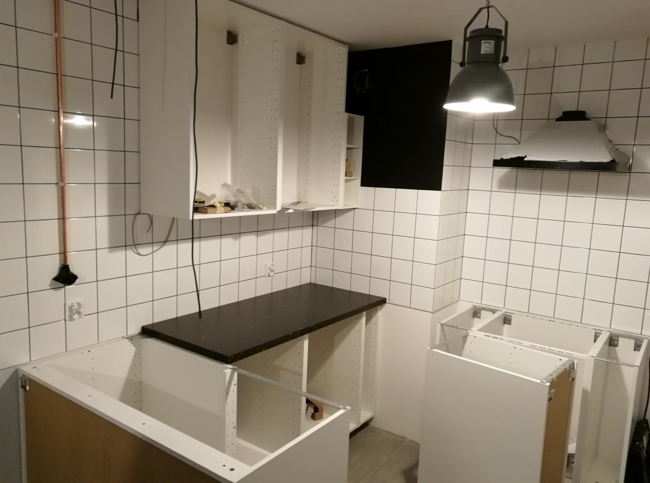Wnętrze kuchni w trakcie montażu: białe szafki wiszące i stojące bez frontów, czarny blat, białe płytki na ścianach, widoczna instalacja elektryczna i wentylacyjna.