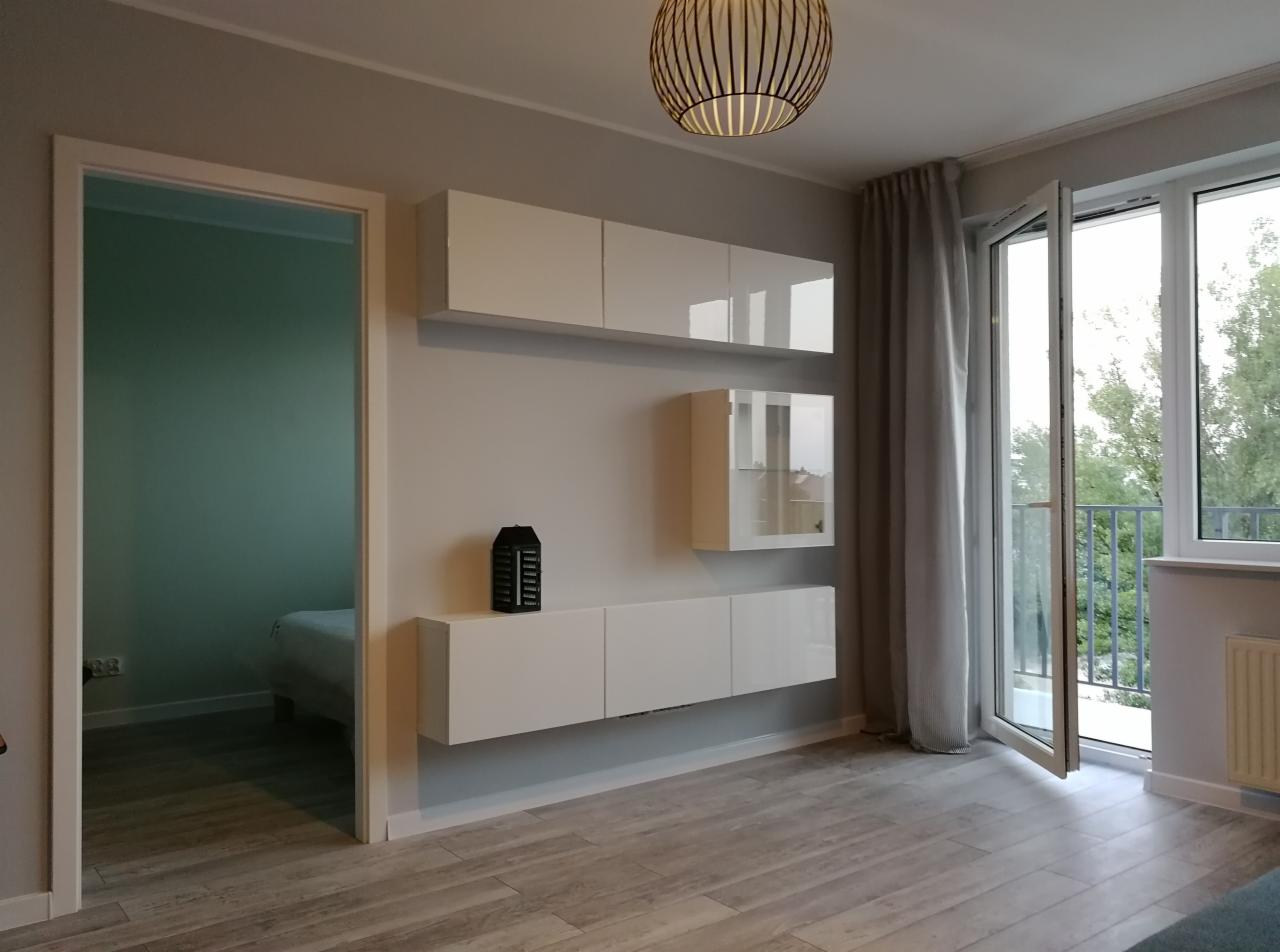 Nowoczesny salon z białymi, podwieszanymi szafkami, drewnianą podłogą i otwartym balkonem z widokiem na zieleń.