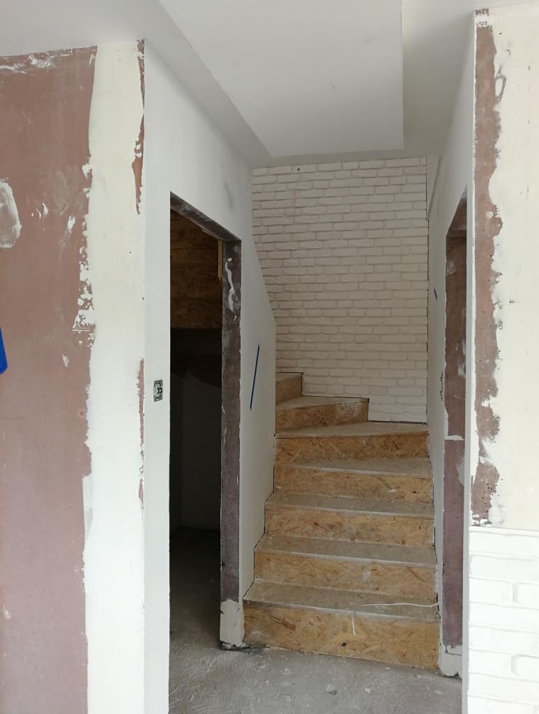 Klatka schodowa w trakcie budowy: surowe drewniane stopnie, ściany w trakcie szpachlowania i malowania, dekoracyjna ściana z białej cegły w tle.