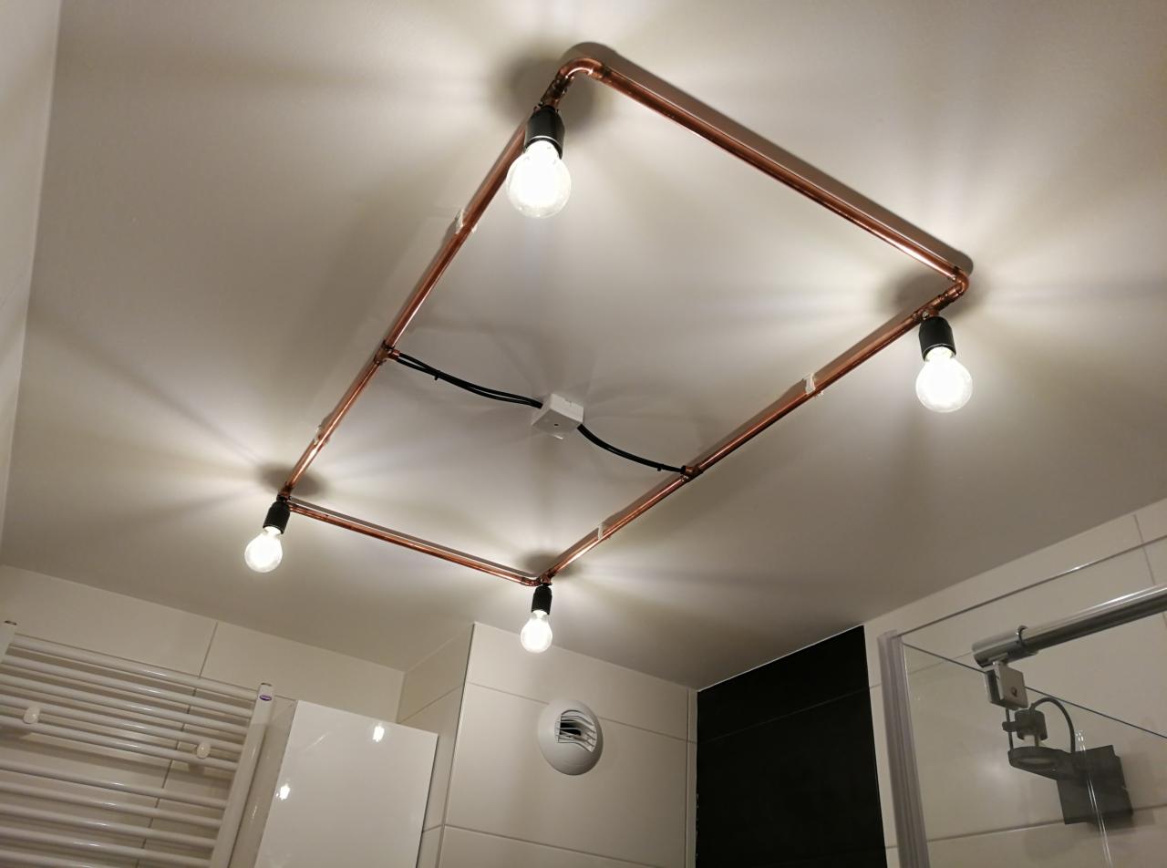 Oryginalna lampa sufitowa z miedzianych rurek i żarówek w stylu industrialnym, zamontowana we wrocławskiej łazience z biało-czarnymi płytkami, widoczny fragment kabiny prysznicowej i grzejnika.