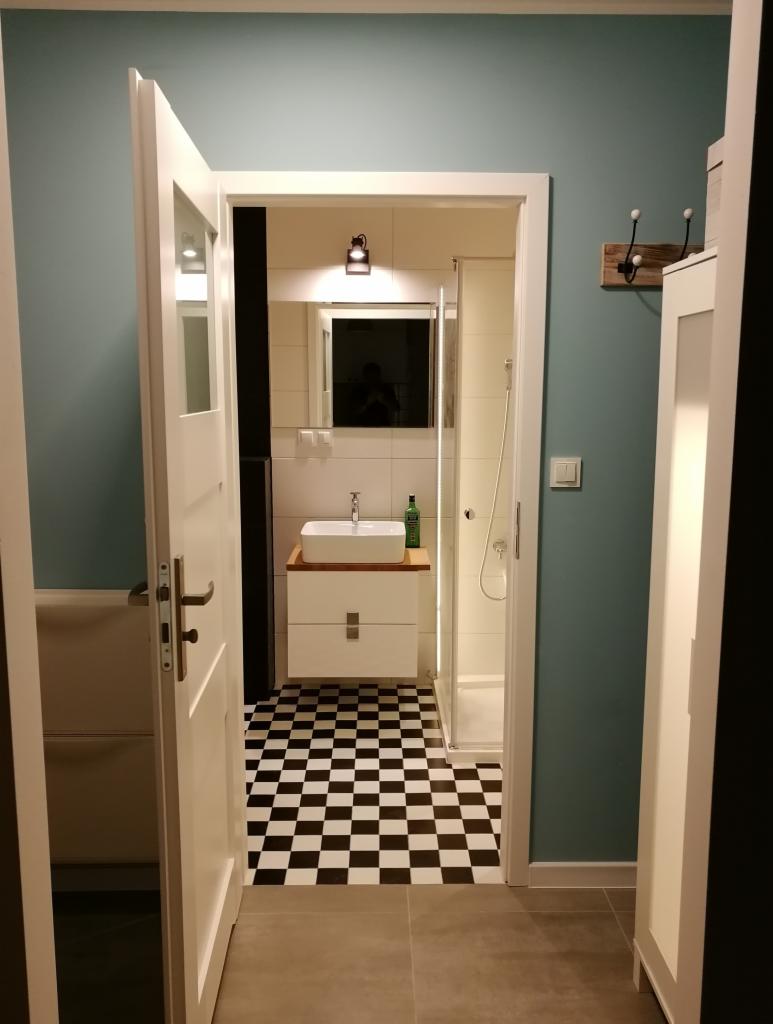 Otwarta biała rama drzwi prowadzi do odnowionej łazienki z białą umywalką na szafce z drewnianym blatem, lustrem, kabiną prysznicową i czarno-białą szachownicą na podłodze.