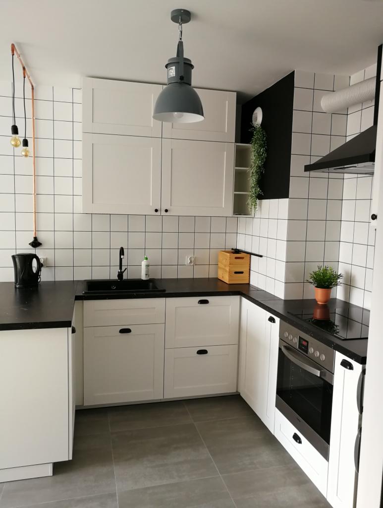 Kuchnia w stylu skandynawskim: białe szafki z czarnymi uchwytami, czarny blat, białe płytki kwadratowe na ścianie, szara podłoga, wisząca lampa w stylu industrialnym i dekoracyjne oświetlenie...